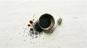 Black seed oil UK — Nigella sativa seeds in an aged ceramic bowl with amber oil drops on pale travertine